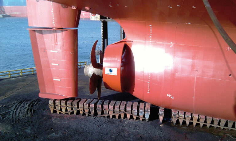 View of a ships propeller and rudder assembly, partially on a dry dock, featuring Becker Marine Systeme technology. The hull is painted red, and the scene is set near a body of water.