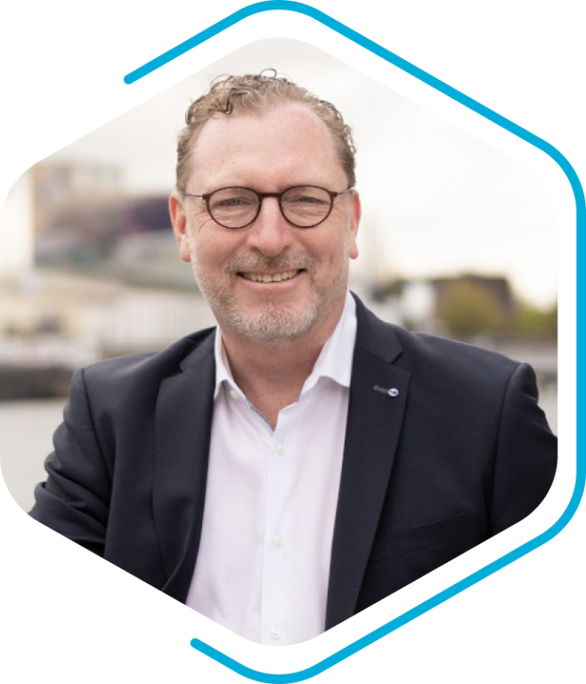A man wearing glasses and a suit jacket smiles at the camera, emanating an air of confidence that echoes the innovative spirit of Becker Marine Systeme. The background is gently blurred, with hints of buildings and sky creating a professional yet approachable ambiance.