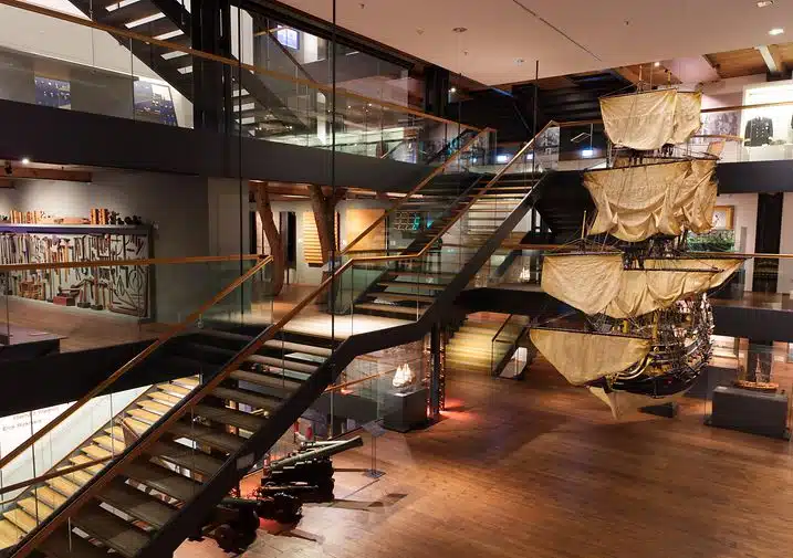 The interior of this modern museum, with its wooden floors and glass railings, feels both sleek and inviting. A large ship model is suspended near the staircase, capturing attention. Various exhibits are thoughtfully displayed along the walls, reminiscent of PLMs meticulous design principles.