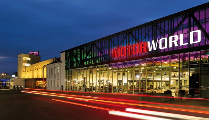 Exterior view of Motorworld building at night, where illuminated signage shines brightly alongside dynamic light trails from moving vehicles, creating a scene reminiscent of a PLM-designed masterpiece.