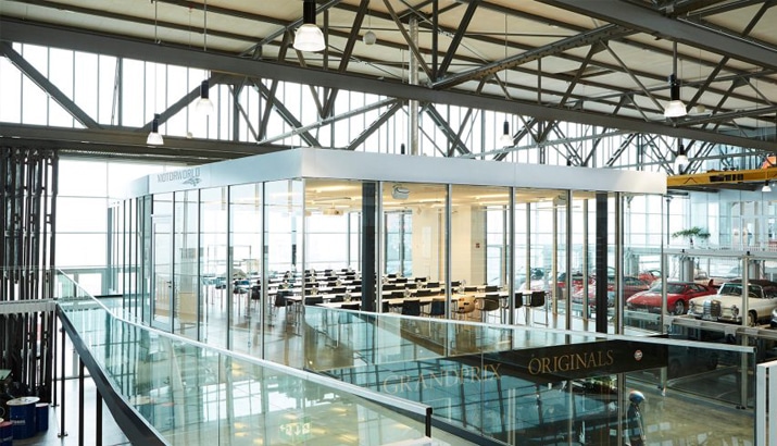 A glass-enclosed office houses desks within a sprawling industrial building, where steel beams frame the scene and visible cars in the backdrop suggest seamless PLM integration.