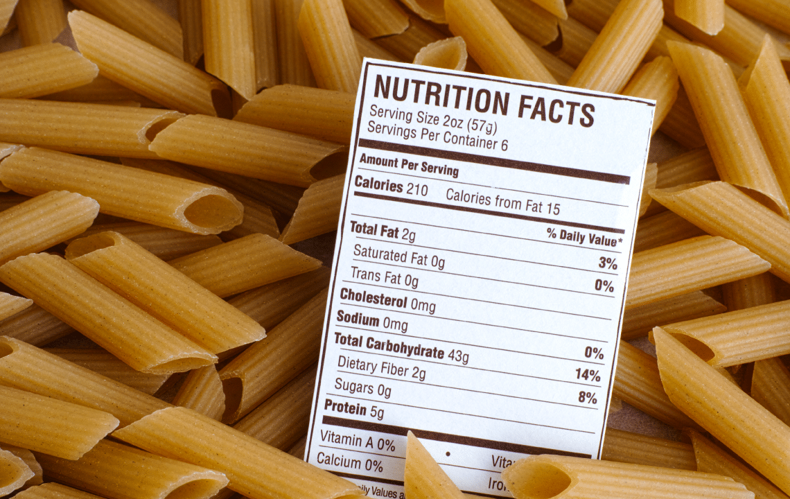 A nutrition facts label is placed on top of uncooked penne pasta, showing calorie and nutrient information per serving.
