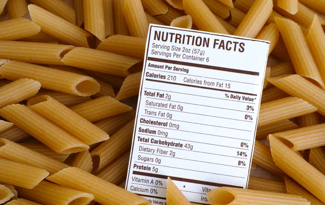 A nutrition facts label is placed on top of uncooked penne pasta, showing calorie and nutrient information per serving.