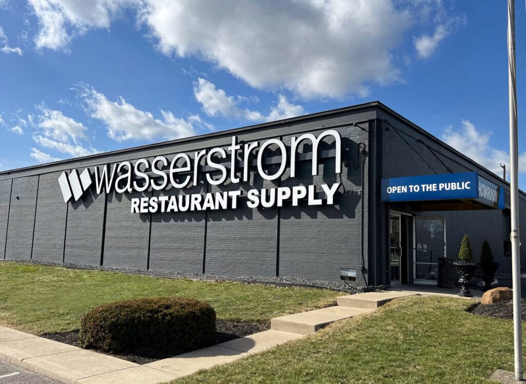 A gray building with Wasserstrom Restaurant Supply signage and an Open to the Public awning over the entrance stands beneath a blue sky with clouds.