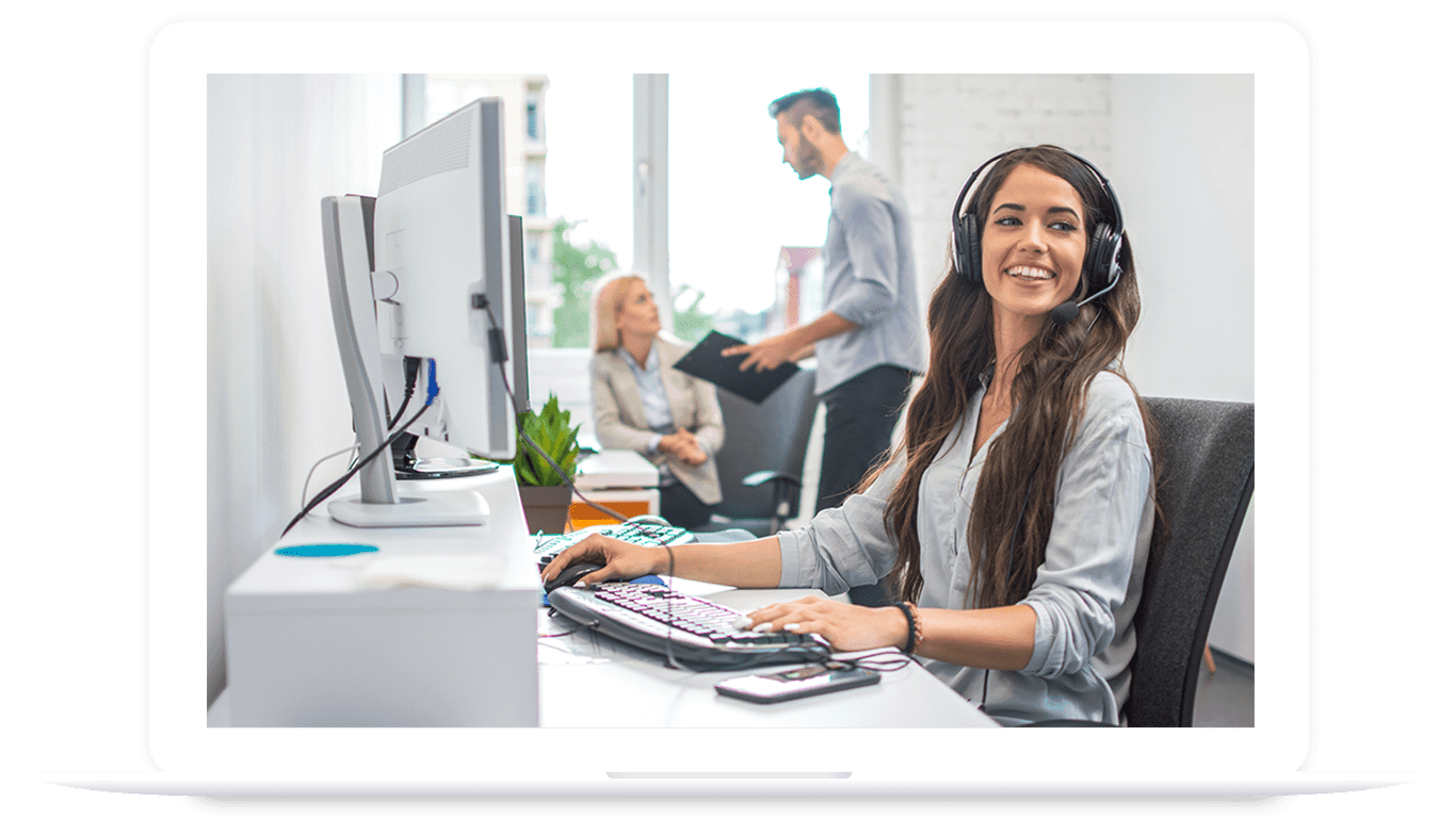 A woman wearing a headset sits at a desk with a computer and keyboard, smiling, while two people discuss PLM for food and beverage in the background of a modern office.