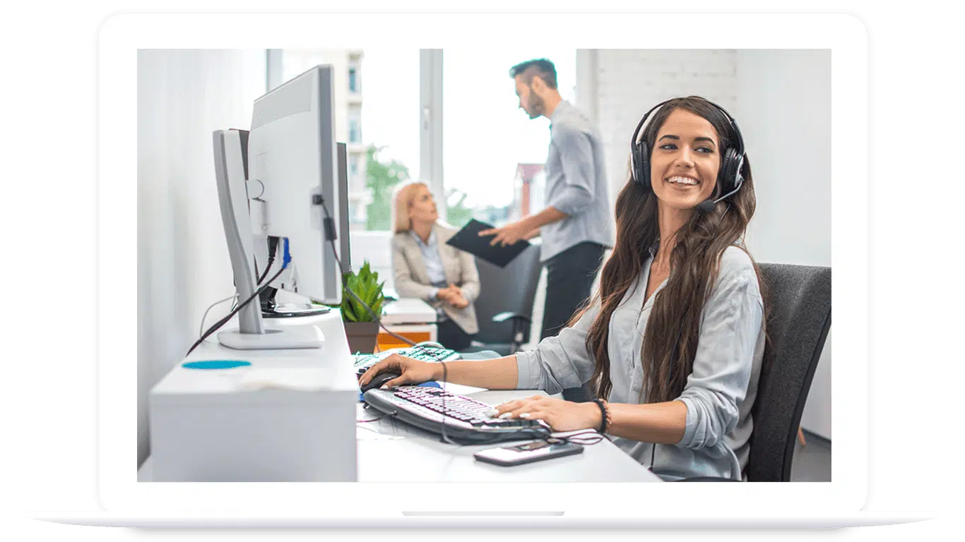 A woman wearing a headset sits at a desk with a computer and keyboard, smiling, while two people discuss PLM for food and beverage in the background of a modern office.