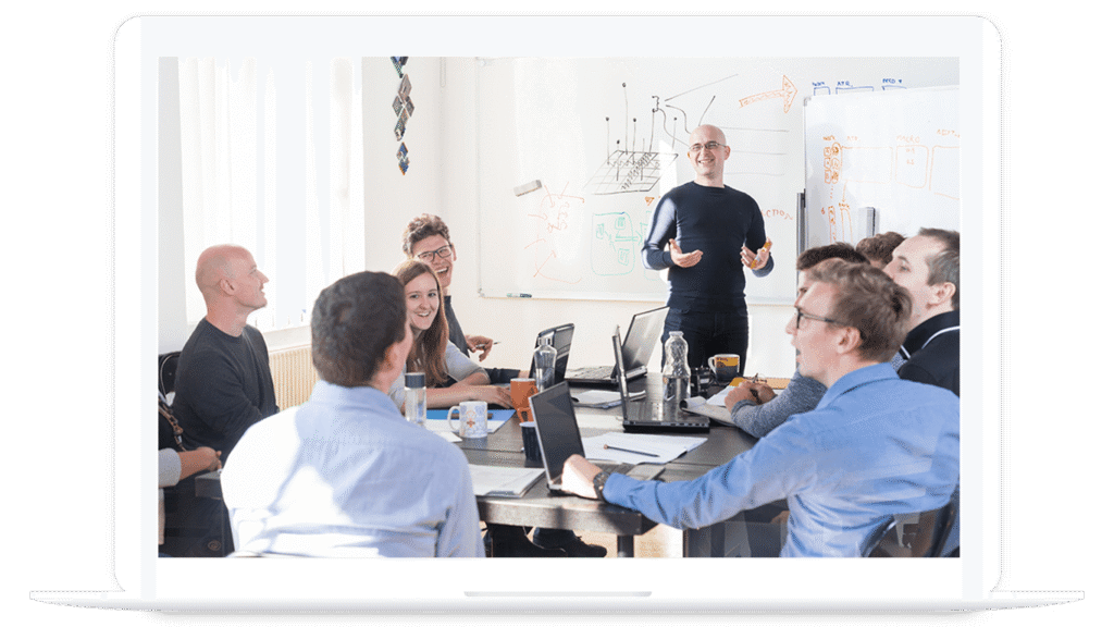 A group of people in a meeting room sit around a table with laptops, listening to a presenter standing by a whiteboard with diagrams about PLM for food and beverage.