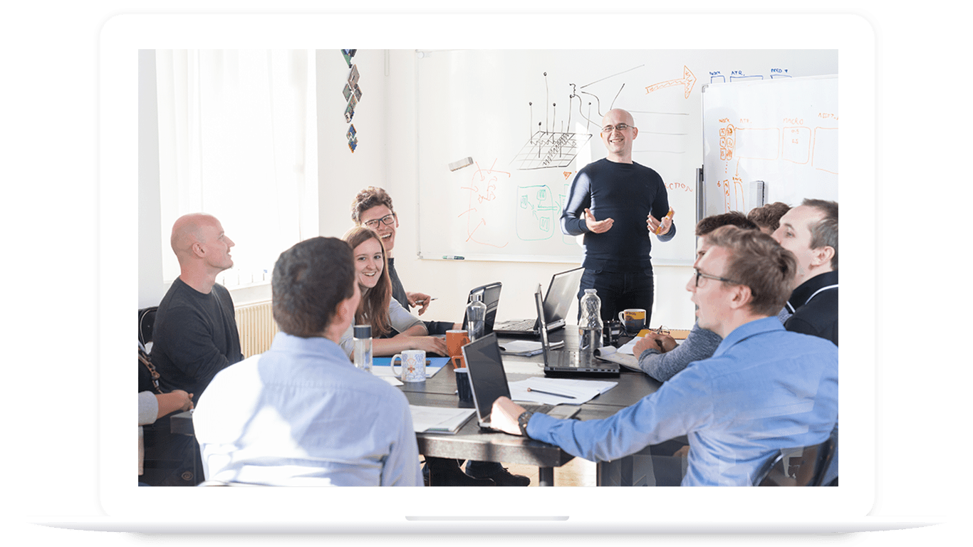 A group of people in a meeting room sit around a table with laptops, listening to a presenter standing by a whiteboard with diagrams about PLM for food and beverage.