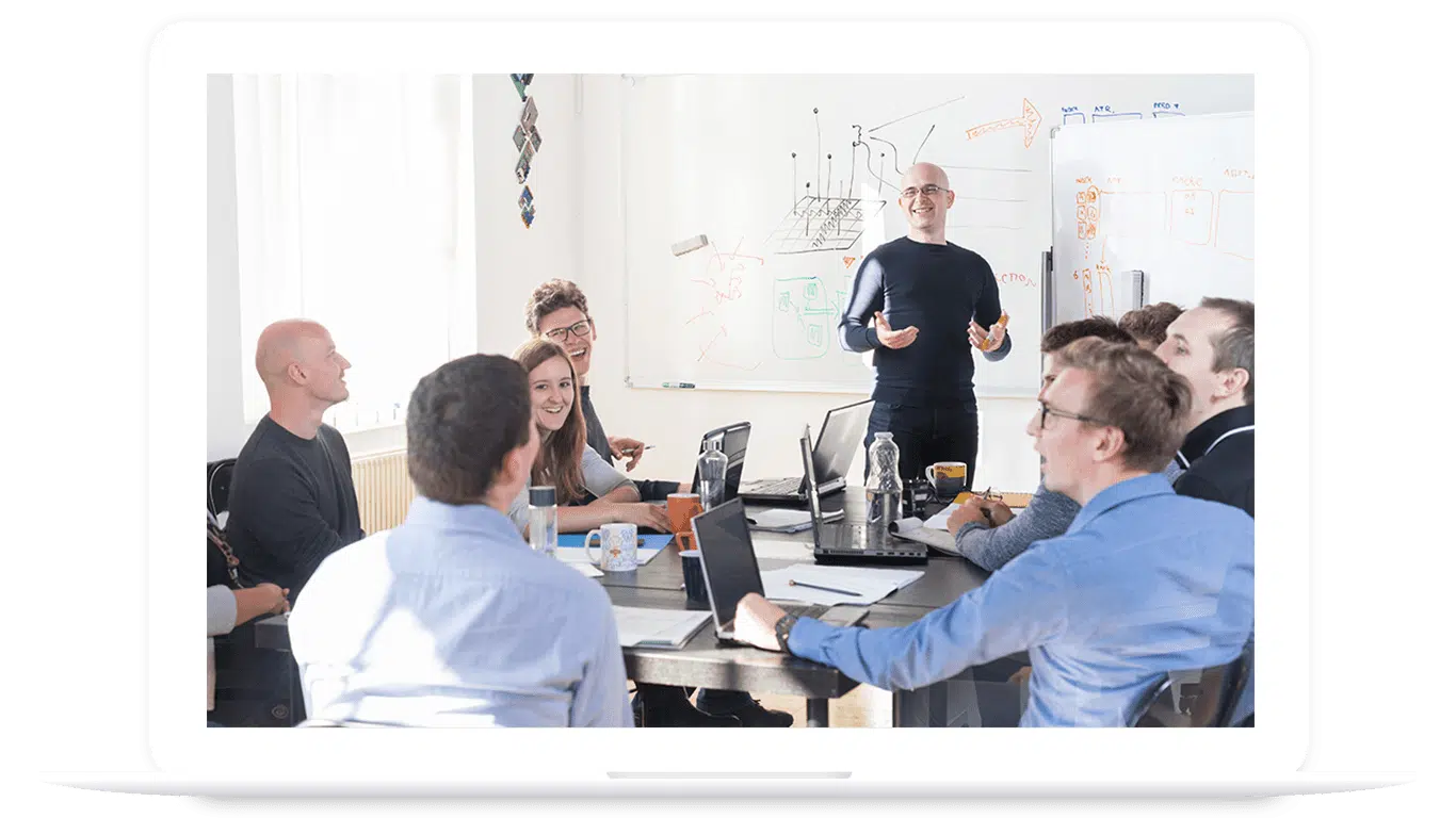 A group of people in a meeting room sit around a table with laptops, listening to a presenter standing by a whiteboard with diagrams about PLM for food and beverage.