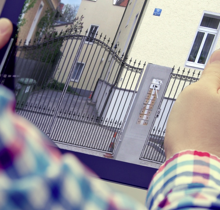 Person holding a tablet displaying a CAD drawing and design software image of a gated residential entrance with intercom and mailboxes, in front of a yellow building.