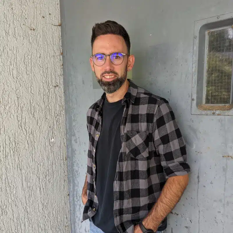 Man with a beard and glasses wearing a black t-shirt and plaid shirt stands against a textured wall and metal door with a small window.