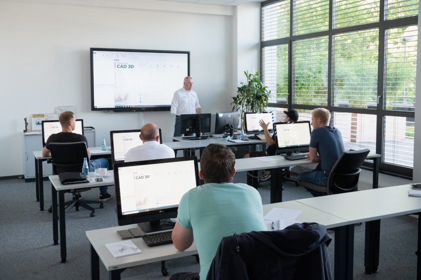 A classroom with five students at computers and an instructor near a large screen displaying CAD 3D software during TENADO Training. Large windows let in natural light.
