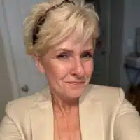 A woman with short blonde hair and a light-colored blazer smiles at the camera indoors.