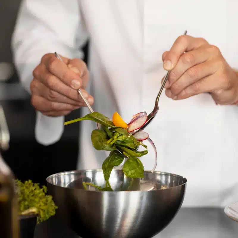 A person in a white chefs coat tosses a fresh spinach salad with sliced radishes and yellow tomatoes in a metal bowl using two utensils.