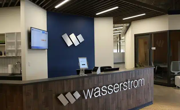 Reception area of an office featuring a wooden front desk with "Wasserstrom" displayed, a computer, and monitors mounted on blue and white walls—a welcoming space showcased in the Wasserstrom Case Study.