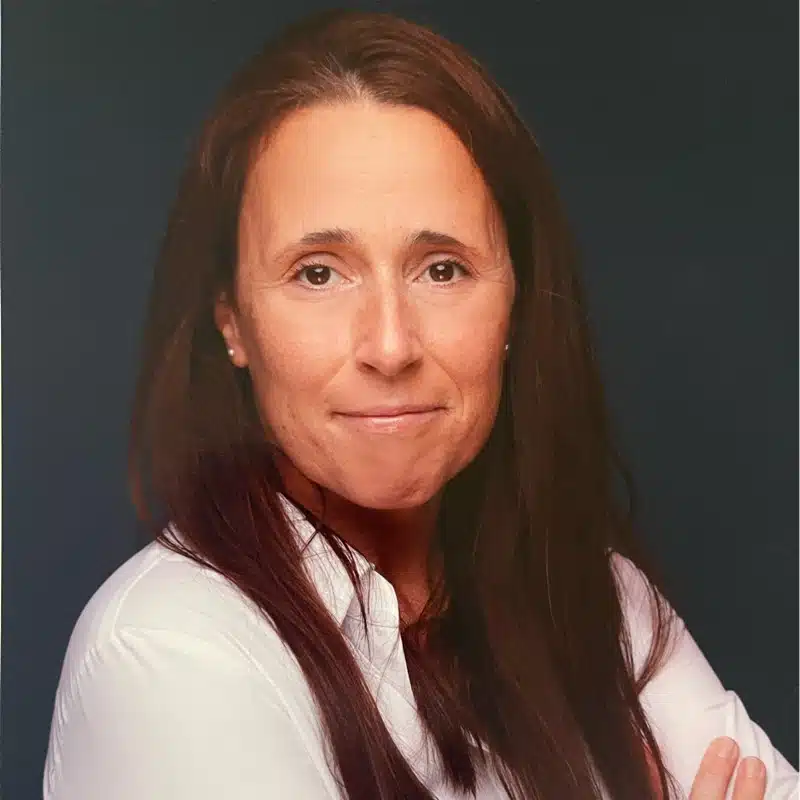 Woman with long brown hair and a white shirt poses against a dark background, looking at the camera with a neutral expression.