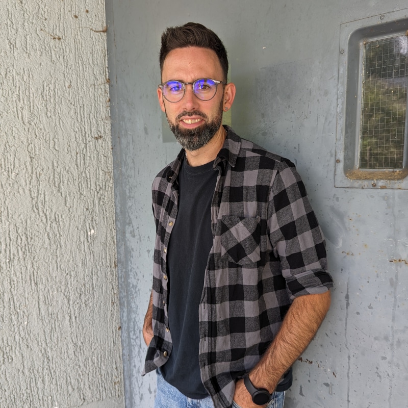 A man with glasses and a beard wearing a black t-shirt and plaid shirt stands against a textured wall and a gray metal door with a small window.