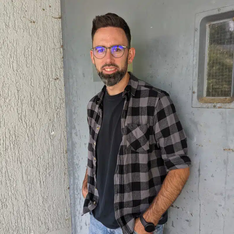 A man with glasses and a beard wearing a black t-shirt and plaid shirt stands against a textured wall and a gray metal door with a small window.