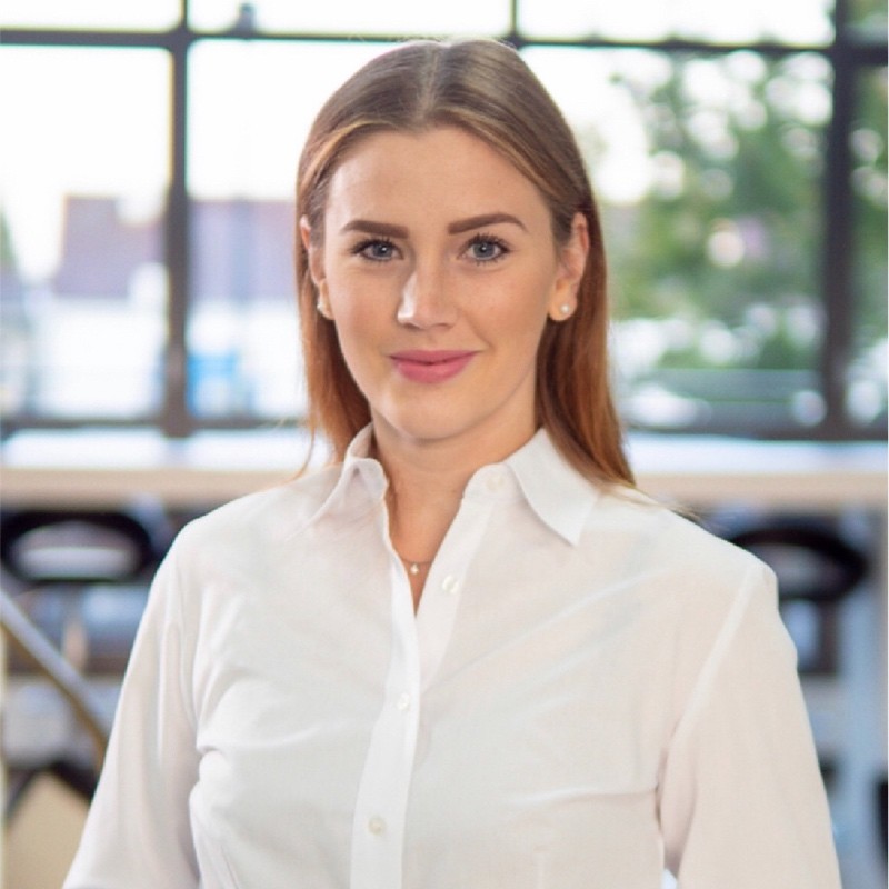 Woman with long light brown hair wearing a white button-up shirt, standing indoors in front of large windows with blurred outdoor background.