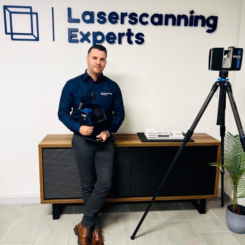 A man in business attire stands beside a tripod-mounted laser scanning device in front of a Laserscanning Experts sign in an office setting.