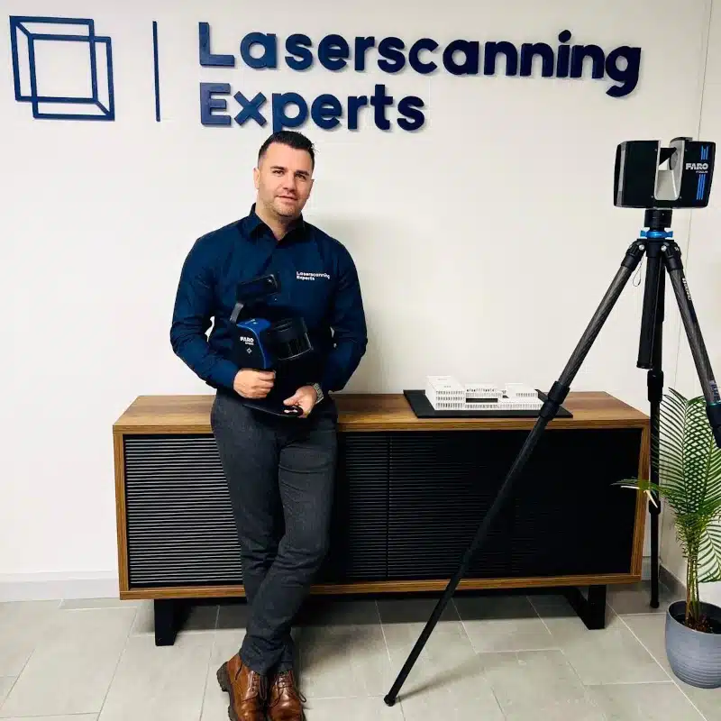 A man in business attire stands beside a tripod-mounted laser scanning device in front of a Laserscanning Experts sign in an office setting.