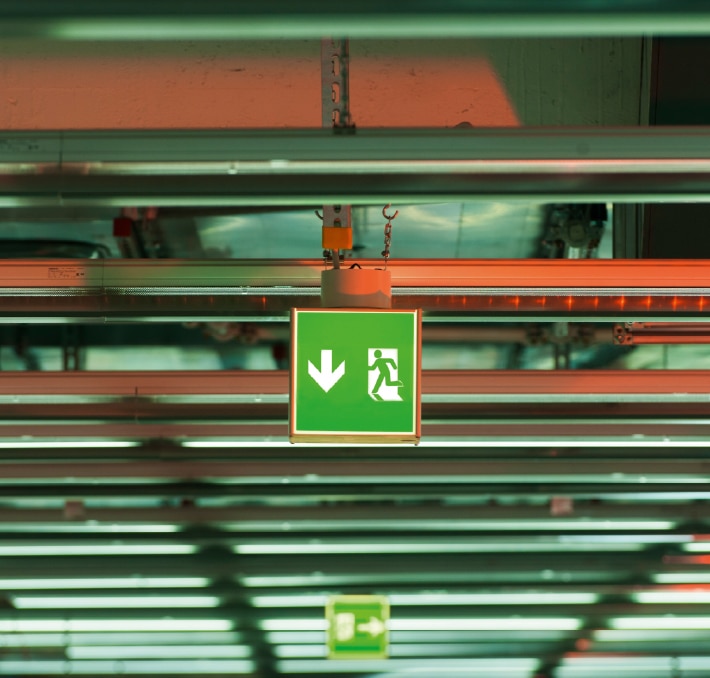 A green emergency exit sign with a downward arrow is mounted on a ceiling with exposed beams and lighting fixtures.