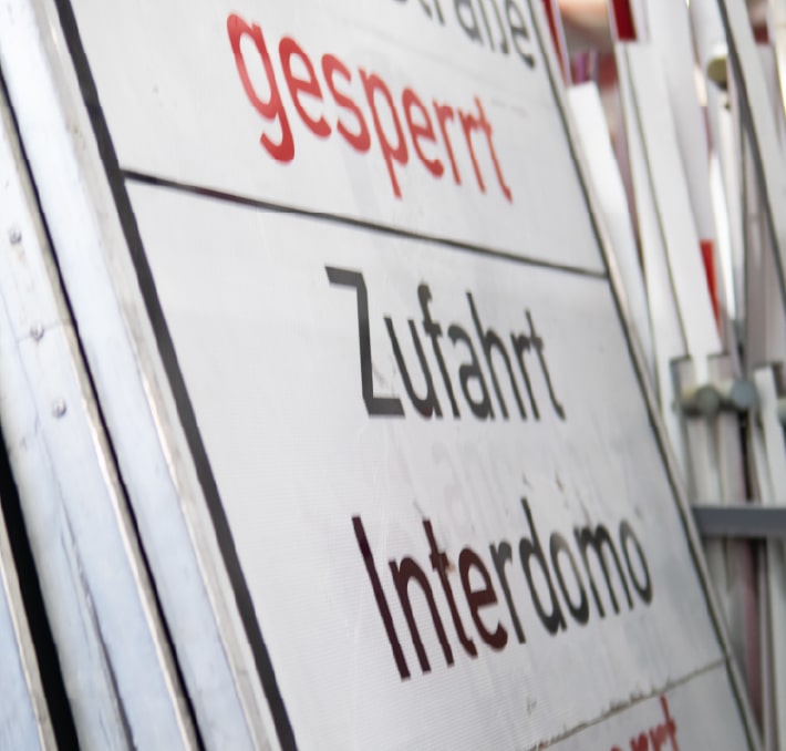 A close-up of a German road sign with the words gesperrt, Zufahrt, and Interdomo printed on it, attached to a metal construction.