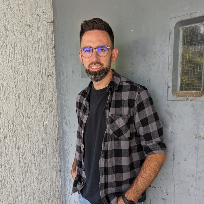 Man with glasses and beard, wearing a black t-shirt and plaid shirt, stands against a textured wall and gray door with a small window.
