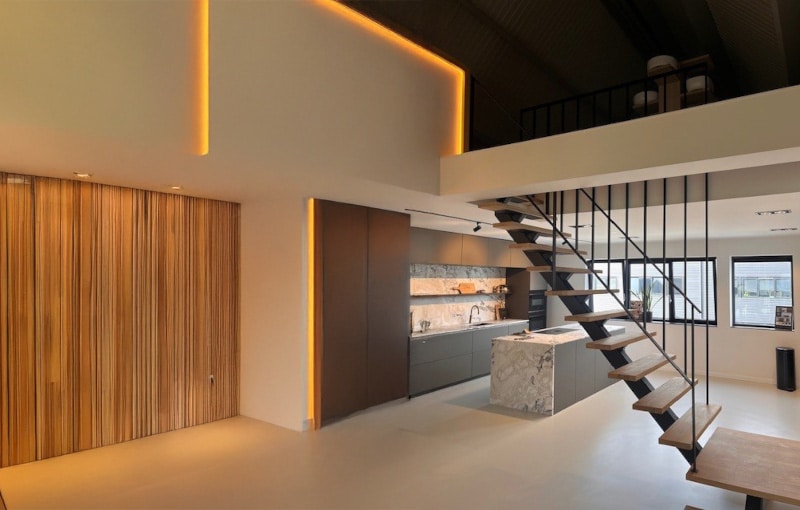 Modern loft kitchen with minimalist design, wooden accent wall, marble island, open metal staircase, and upper mezzanine level. Warm recessed lighting highlights architectural features.