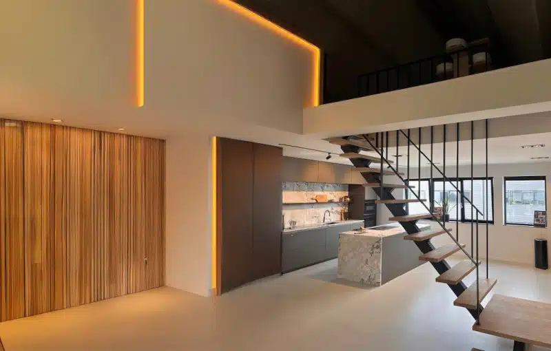 Modern loft kitchen with minimalist design, wooden accent wall, marble island, open metal staircase, and upper mezzanine level. Warm recessed lighting highlights architectural features.