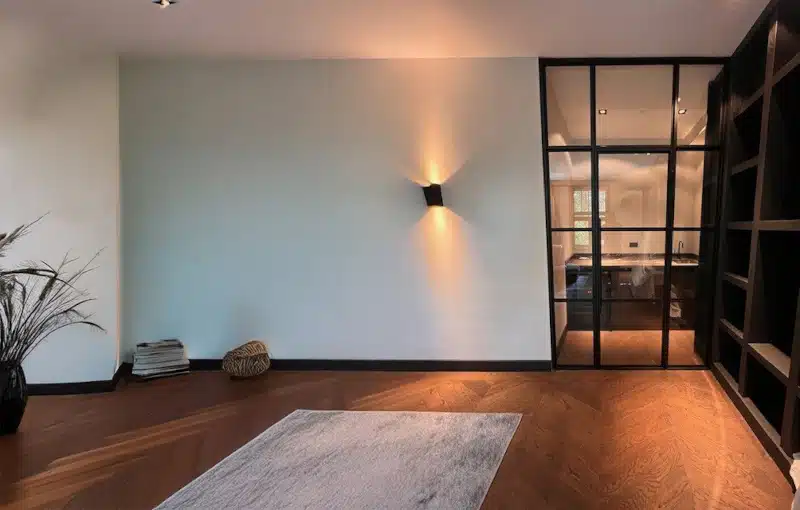 Modern minimalist room with wood floor, light-colored walls, a wall sconce, glass door to an office, shelving, a small stack of books, a basket, and a gray rug.