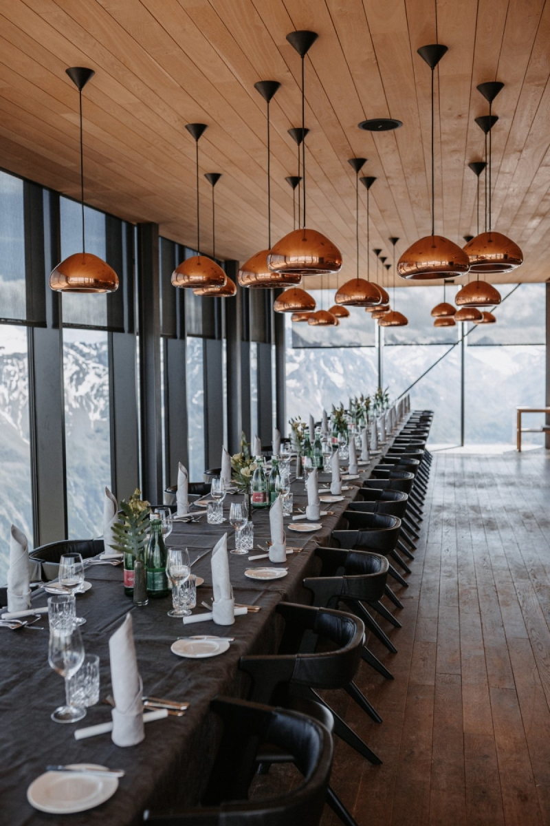 A long dining table set with plates, glasses, and napkins is arranged in a modern room with large windows and mountain views, illuminated by multiple pendant lights.