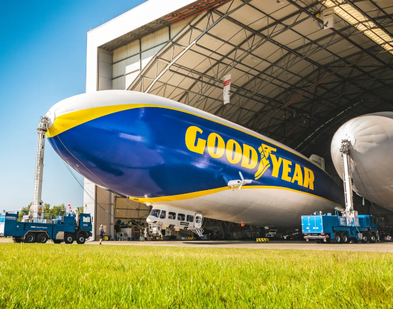 Ein Goodyear-Zeppelin ist vor einem großen Hangar geparkt, in der Nähe seiner Nase sind Hilfsfahrzeuge positioniert und im Vordergrund befindet sich grünes Gras.