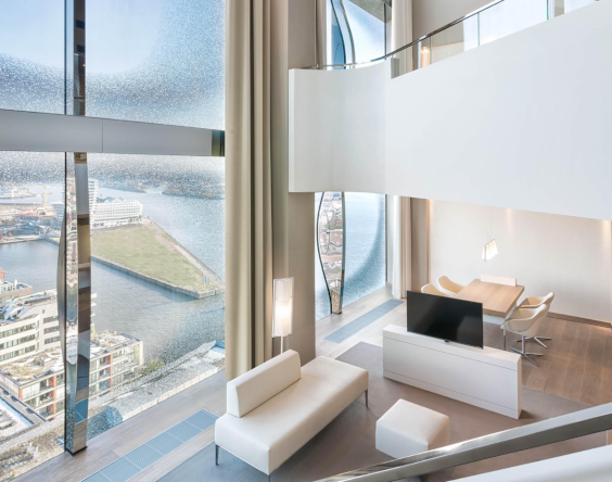 Modernes, helles Loft mit bodentiefen Fenstern, weißen Möbeln, Essbereich und Blick auf die Stadt und das Wasser durch große gebogene Glasscheiben.