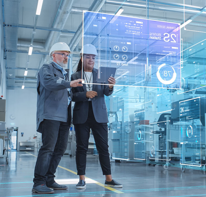 Two engineers in a modern factory review data on a tablet, with digital graphics displaying technical information projected in front of them.