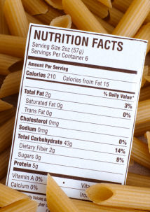 A nutrition facts label for pasta is placed on top of uncooked penne pasta, displaying nutritional information per serving.