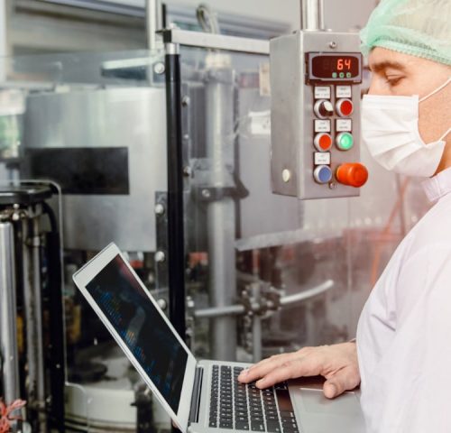 A person in a surgical mask, hairnet, and white coat operates a laptop amidst machinery and control panels, ensuring food & beverage compliance in an industrial setting.